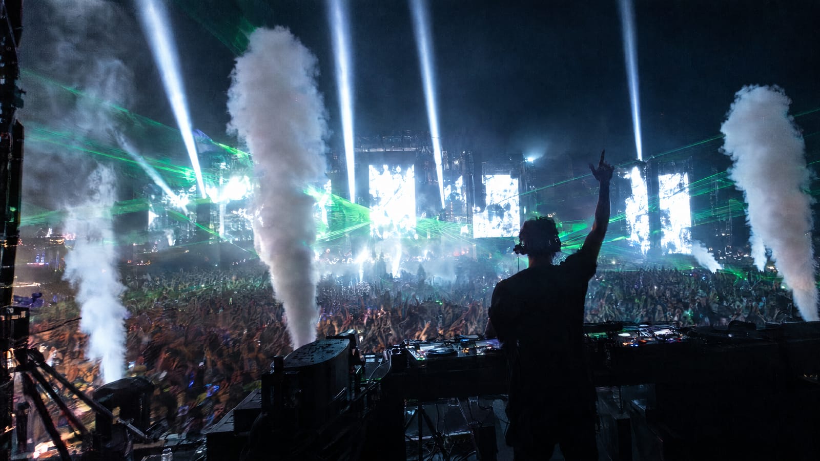 The drop at a night festival seen from behind the DJ: LED walls flash white while moving-head beams, green lasers and CO2 jets fire at the same moment