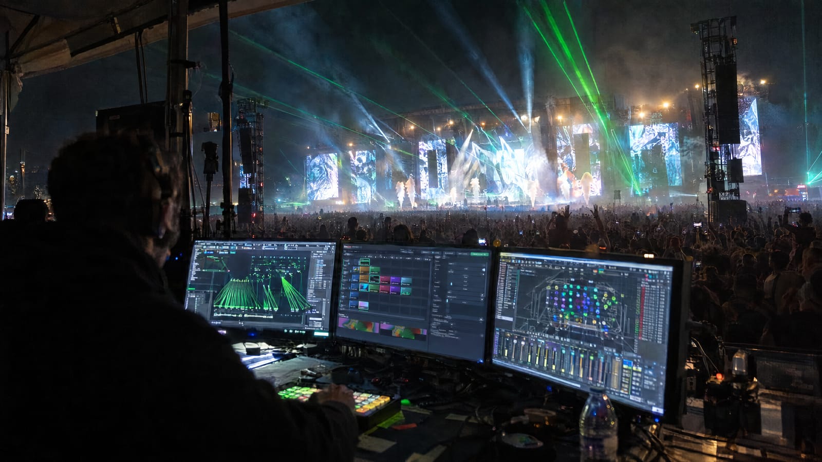 Front-of-house tent at a night festival: a show operator at three screens running laser control, a VJ clip grid and a DMX lighting plan, with the main stage's LED video walls, green laser beams, moving heads and CO2 jets over the crowd