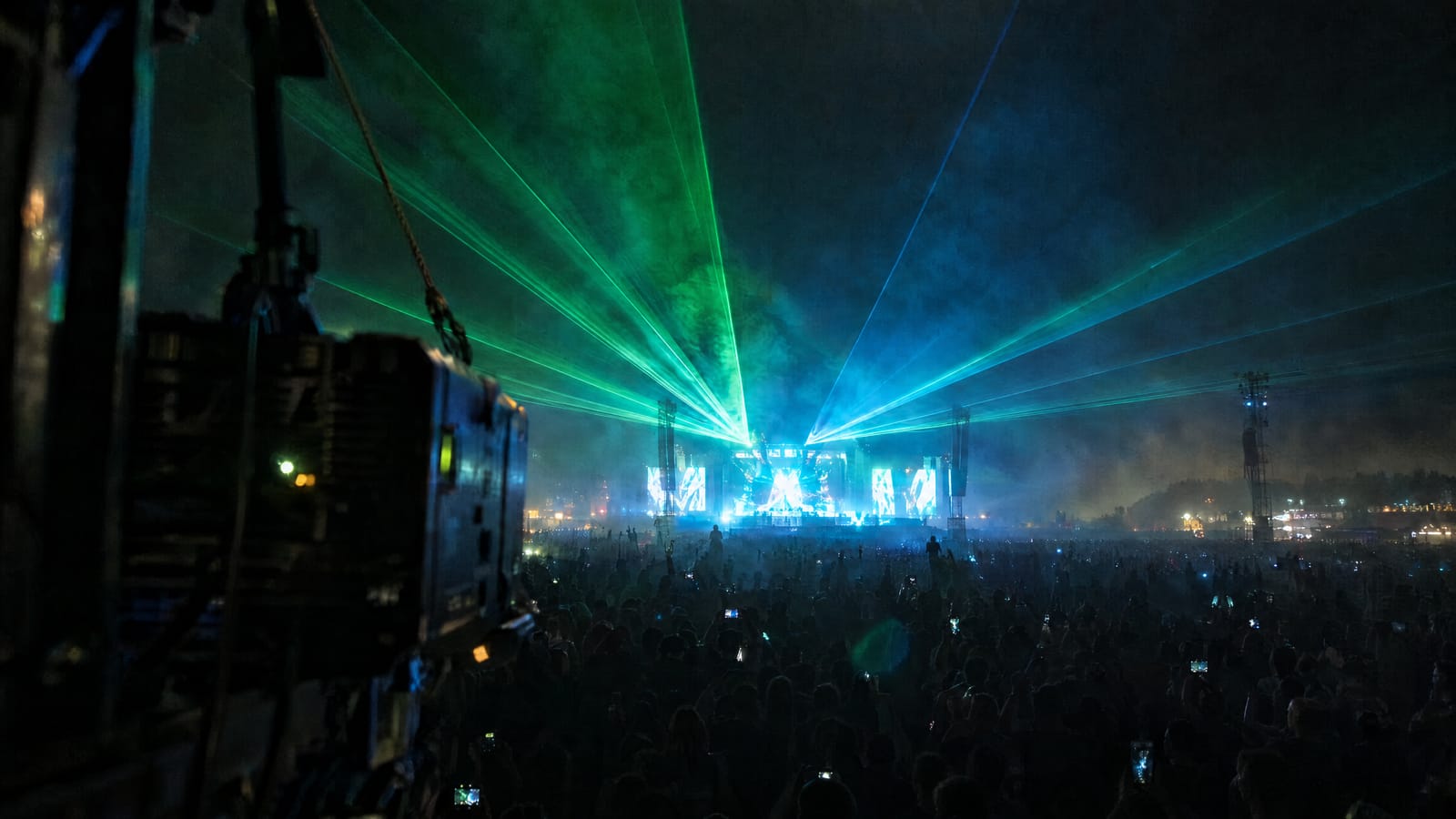 Green and cyan laser fans spreading above a festival crowd from a laser projector on a truss tower, with the main stage far behind
