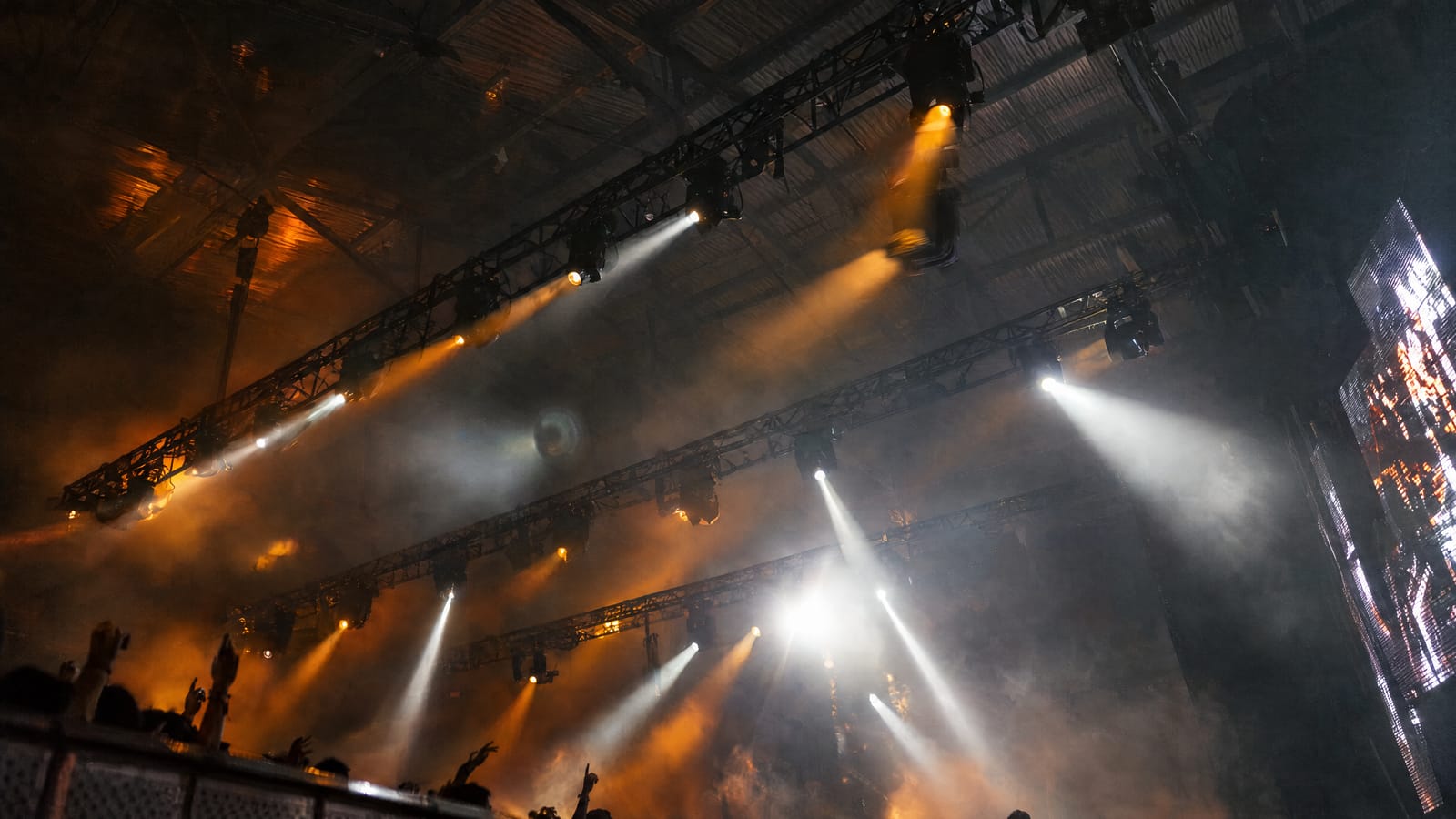 Moving-head lights on truss throwing white and amber beams through haze over a warehouse crowd, the kind of rig a DMX lighting console busks live