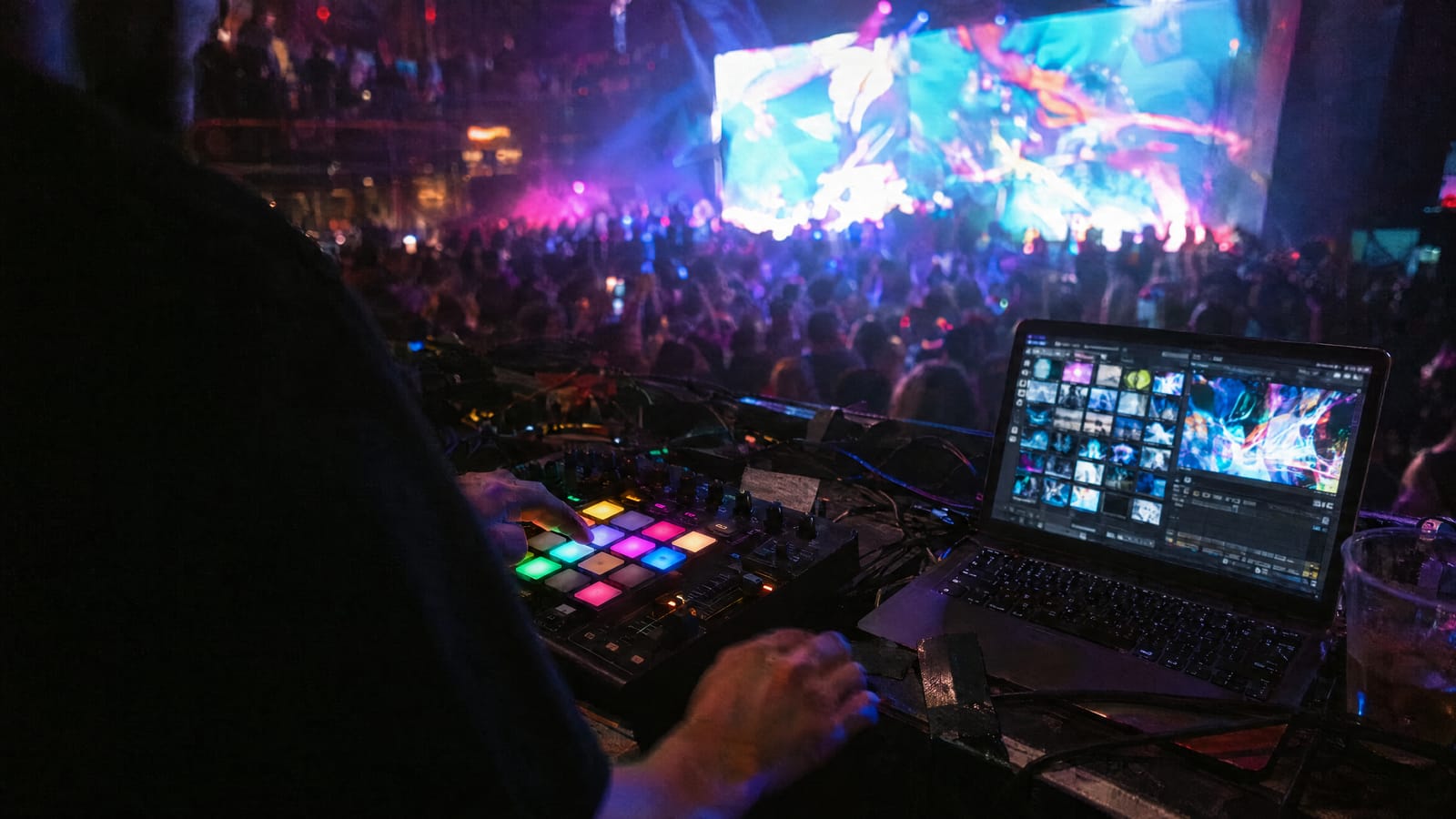 VJ hands on a lit MIDI pad controller beside a laptop running a video clip grid, with an LED wall and a club crowd behind